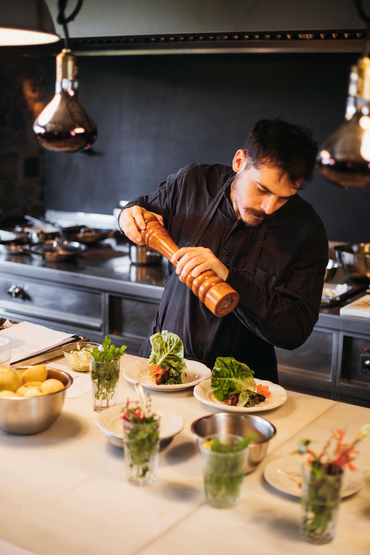 A chef seasoning small plates and glasses of herbs at the kitchen pass
