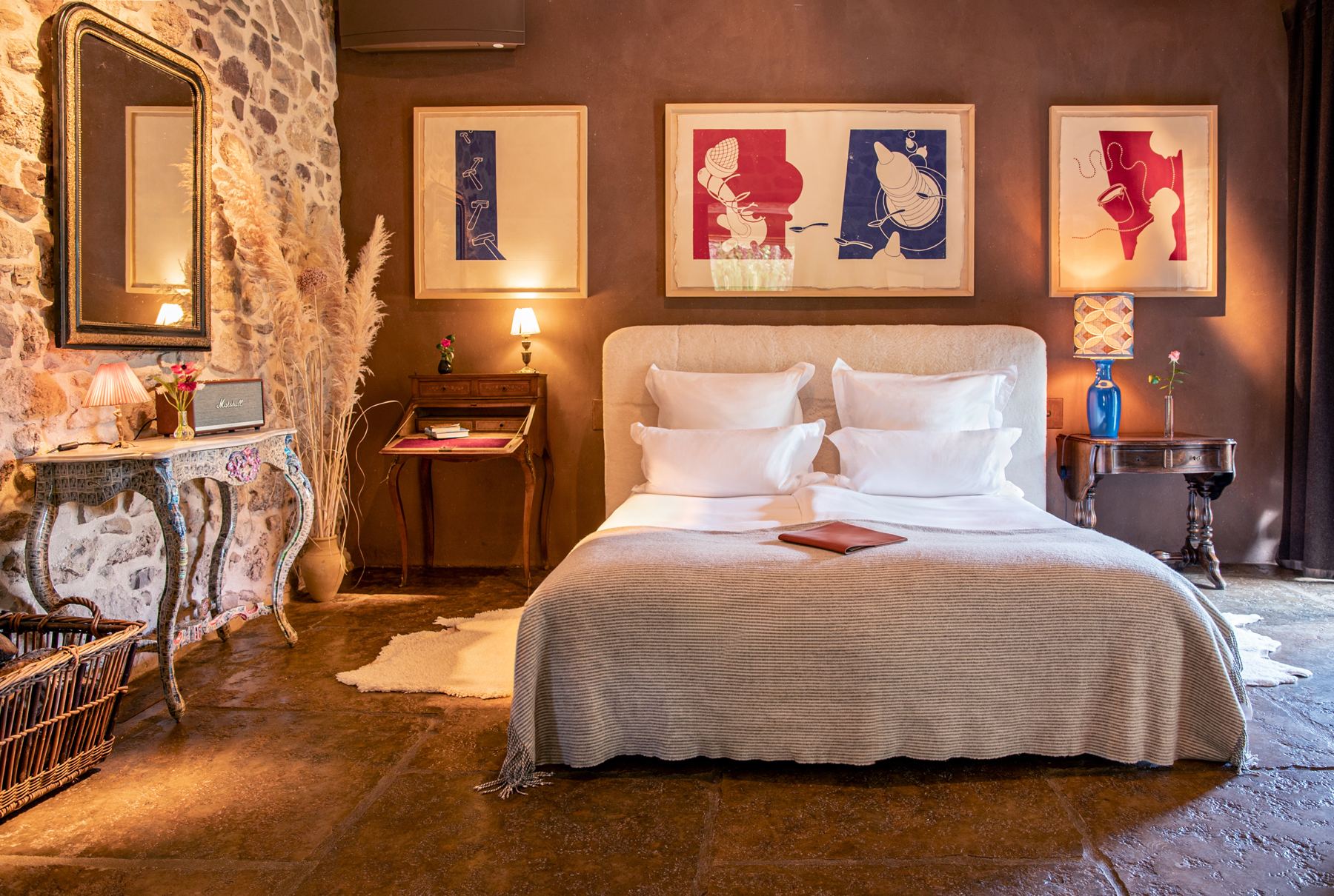 A stone-walled suite with an upholstered bed, antique side tables, pampas grass and three framed red and blue prints