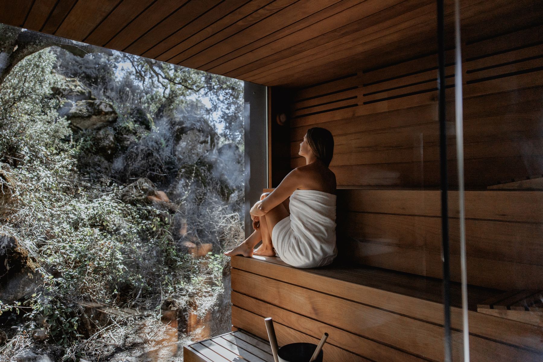 A woman in a towel sitting in a wooden sauna with a glass wall onto oaks and rocks