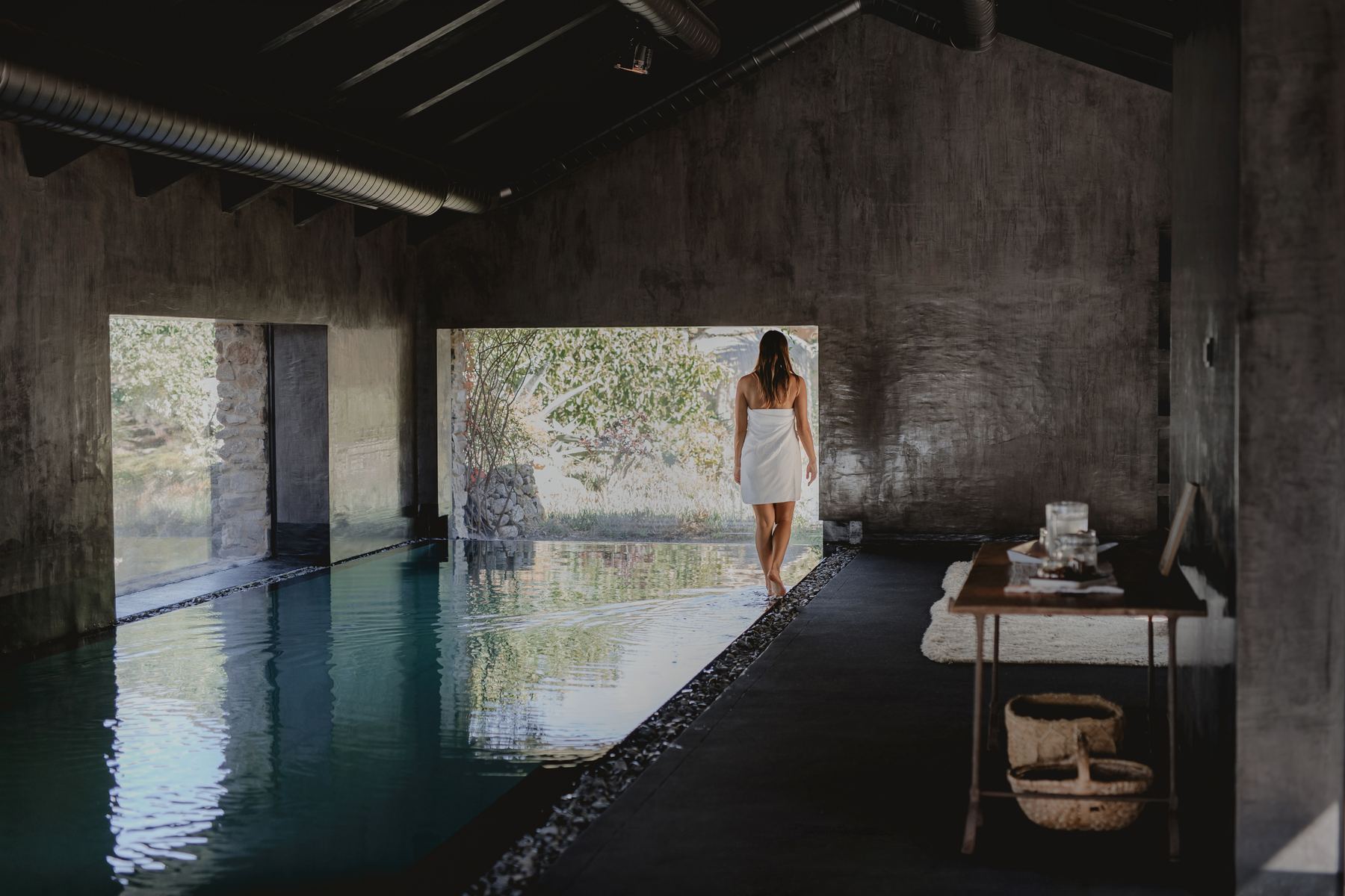 A long dark indoor pool in a stone hall, a woman standing at the far window
