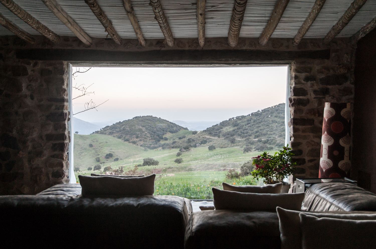 A wide stone-framed window in a dark lounge looking out on green hills at dusk