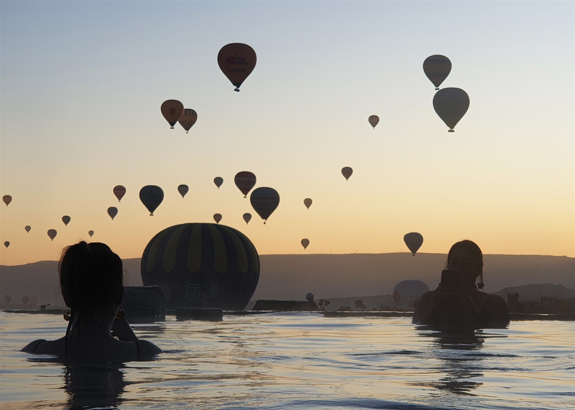 Guests silhouetted in the infinity pool at Museum Hotel as hot air balloons fill the sunrise sky over Cappadocia