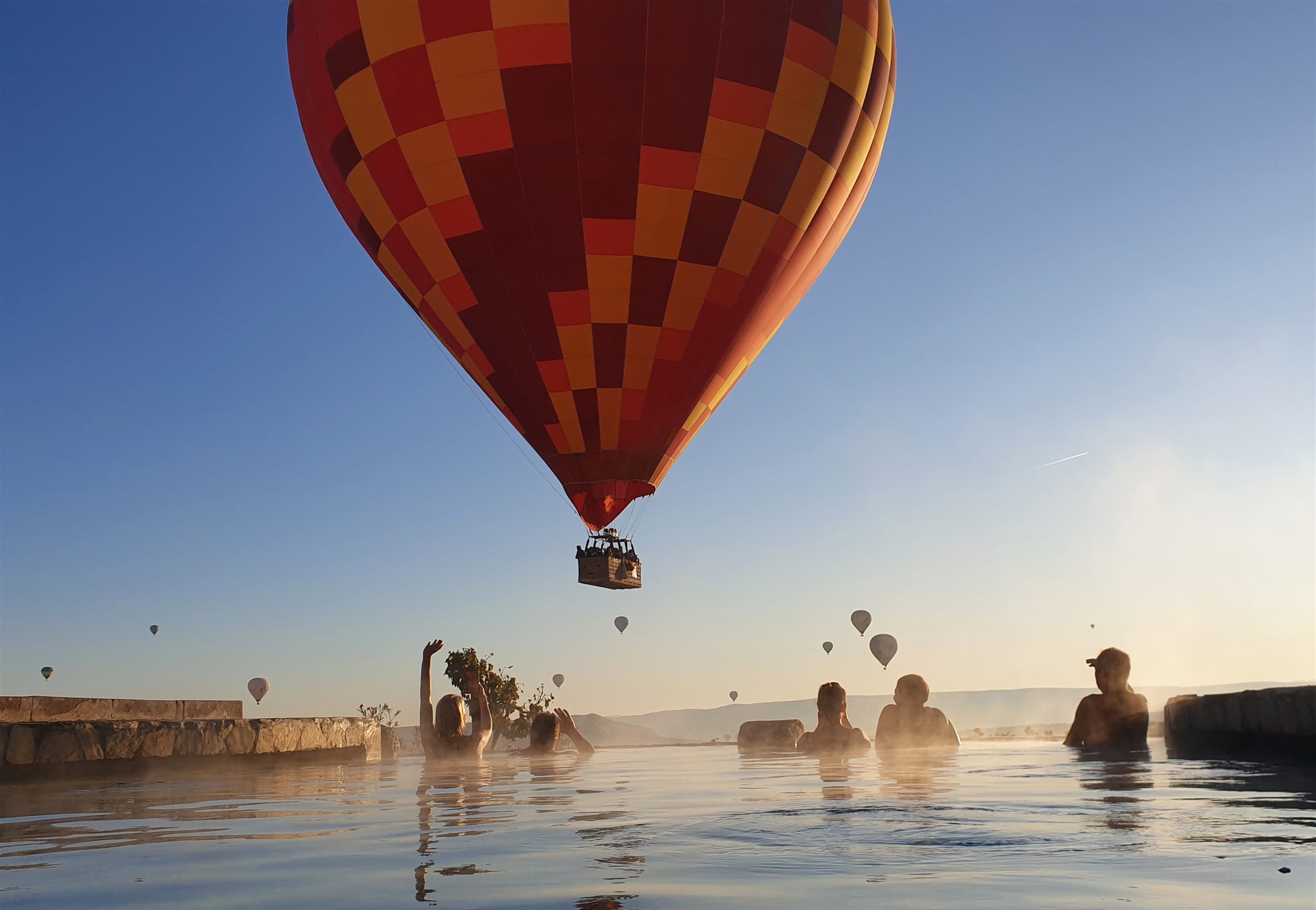 Red and yellow hot air balloon floating low over the pool at Museum Hotel, Cappadocia, with swimmers below