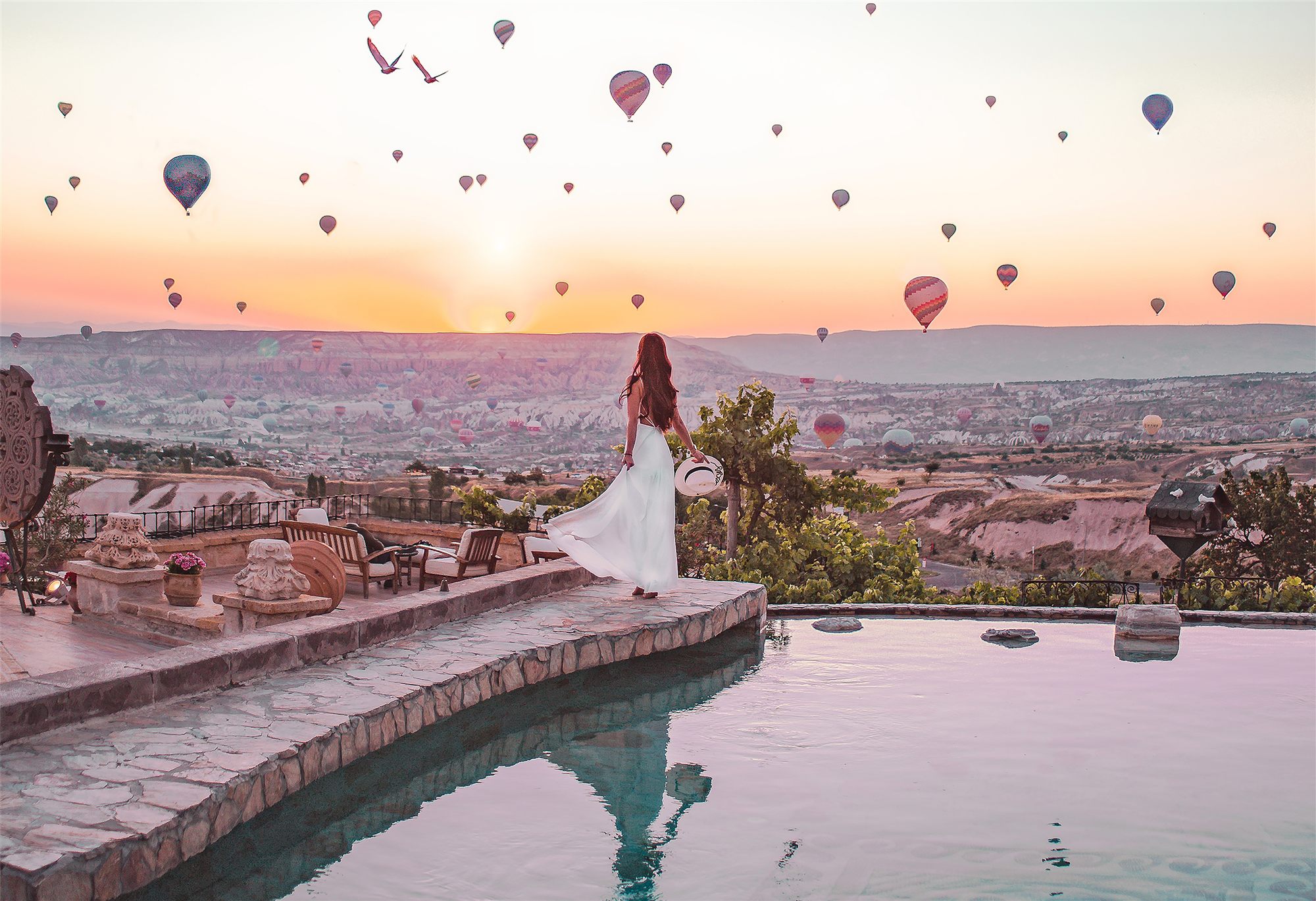 Woman in white standing at the edge of the pool at Museum Hotel, Uçhisar, as hot air balloons rise over Cappadocia at sunrise