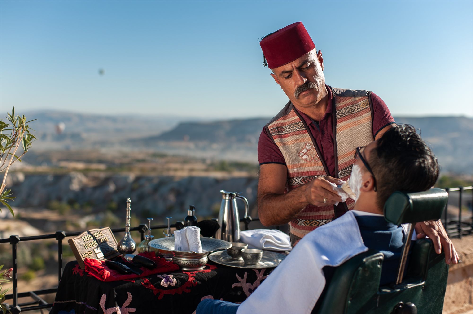 Barber in a red fez giving a traditional Turkish shave on a terrace overlooking Cappadocia