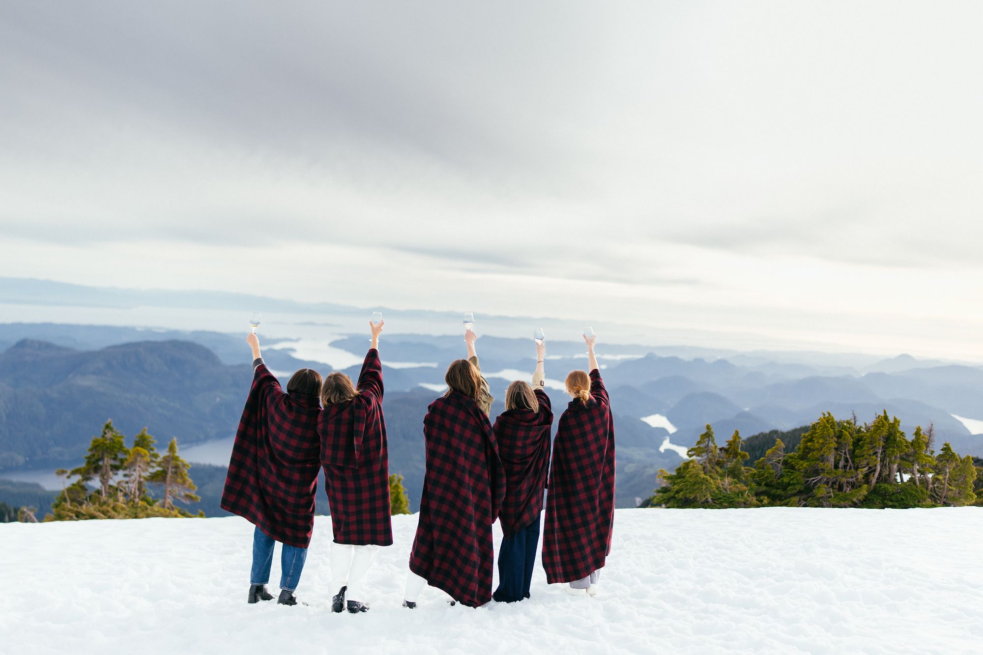 Guests wrapped in blankets raising a toast on a snowy mountaintop after a helicopter flight from Nimmo Bay
