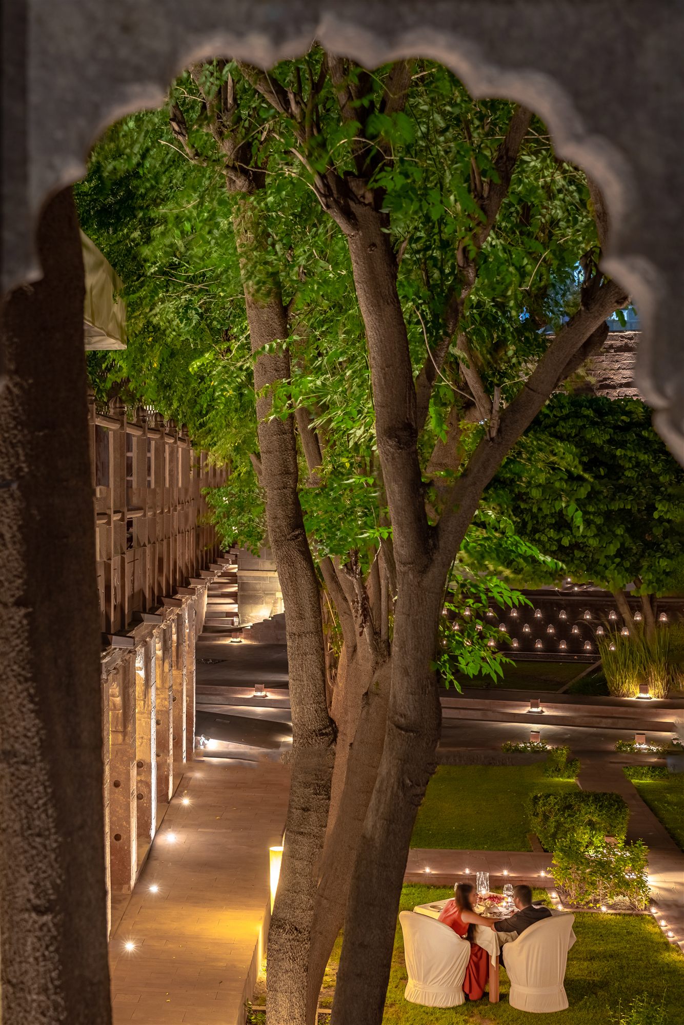 Couple dining by lantern light in the garden beneath a tree, framed by a sandstone arch at RAAS Jodhpur