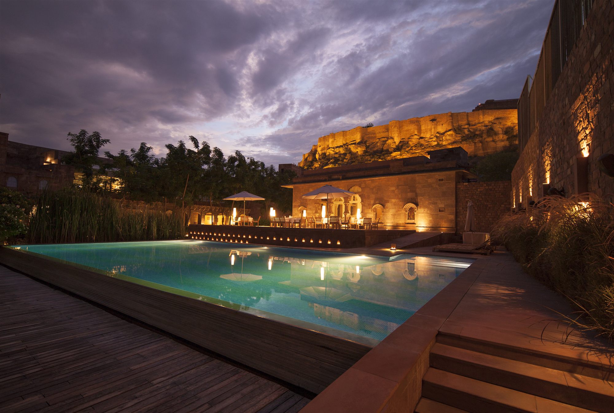 Swimming pool at RAAS Jodhpur at dusk under a dramatic sky, with Mehrangarh Fort illuminated above