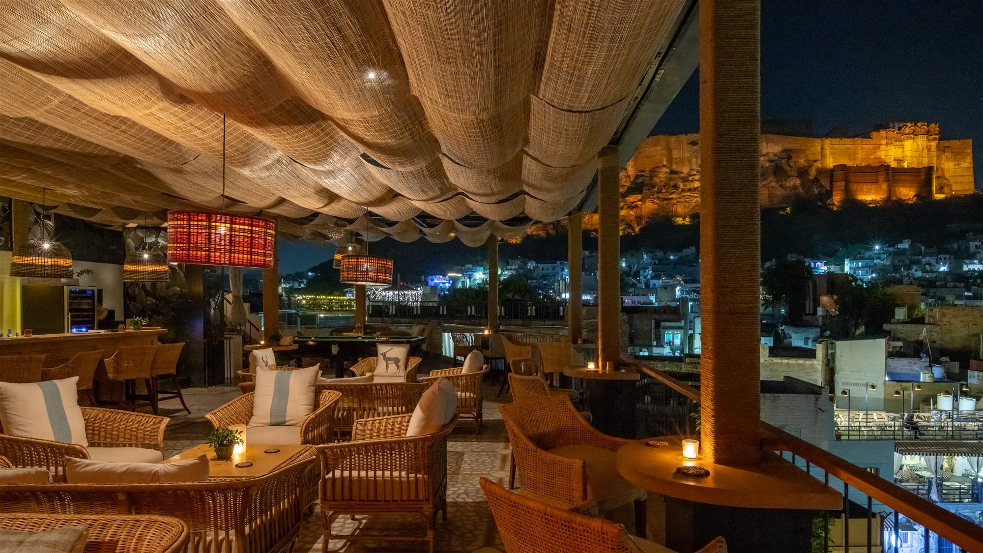The Stepwell Hotel Rooftop Restaurant in Jodhpur at night with woven lanterns, rattan seating and Mehrangarh Fort lit in the distance
