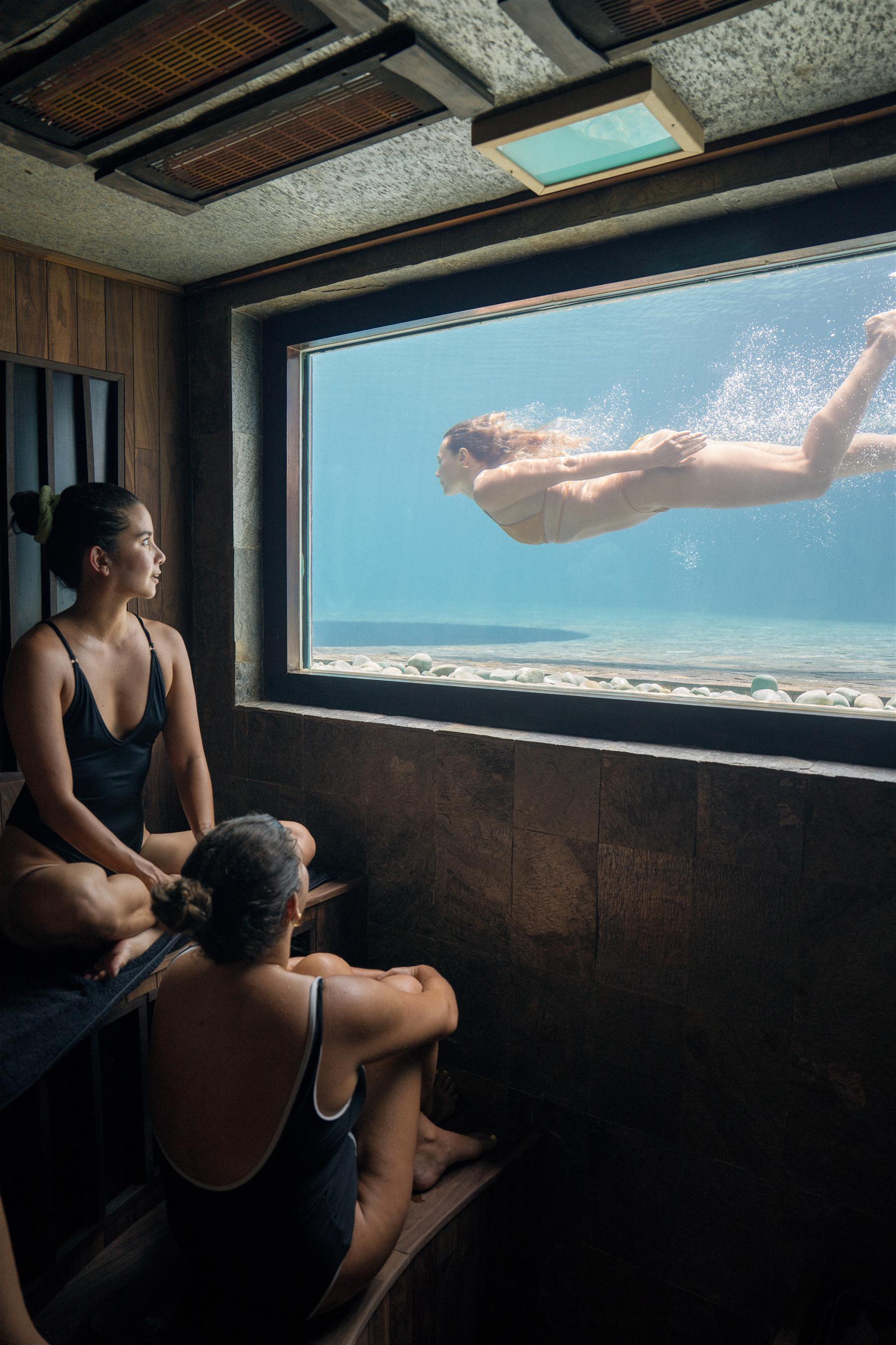 Two women in a wood-paneled sauna watch a swimmer through an underwater pool window at The Seed, Bali