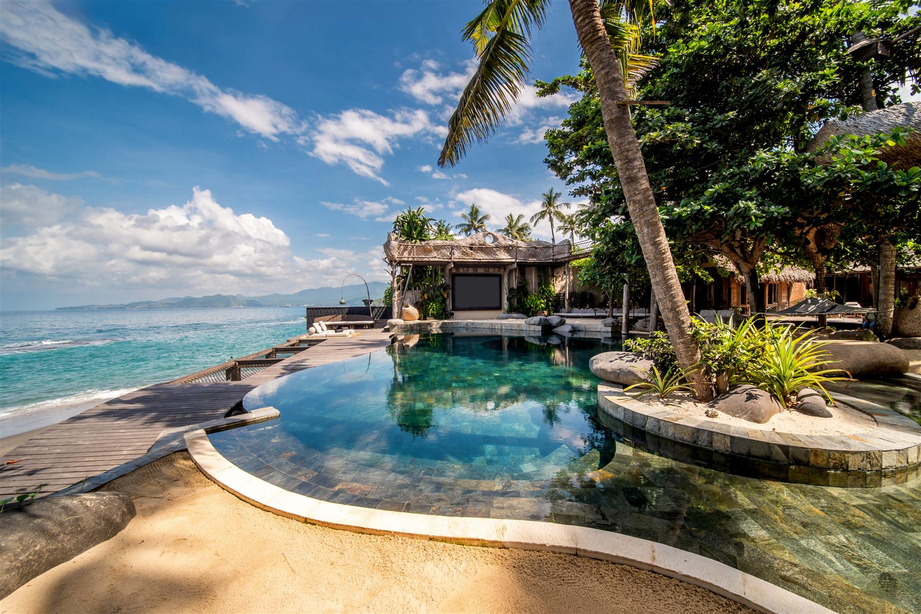 Curved infinity pool at The Seed resort overlooking the ocean in Candidasa, Bali, with palm trees and thatched villas