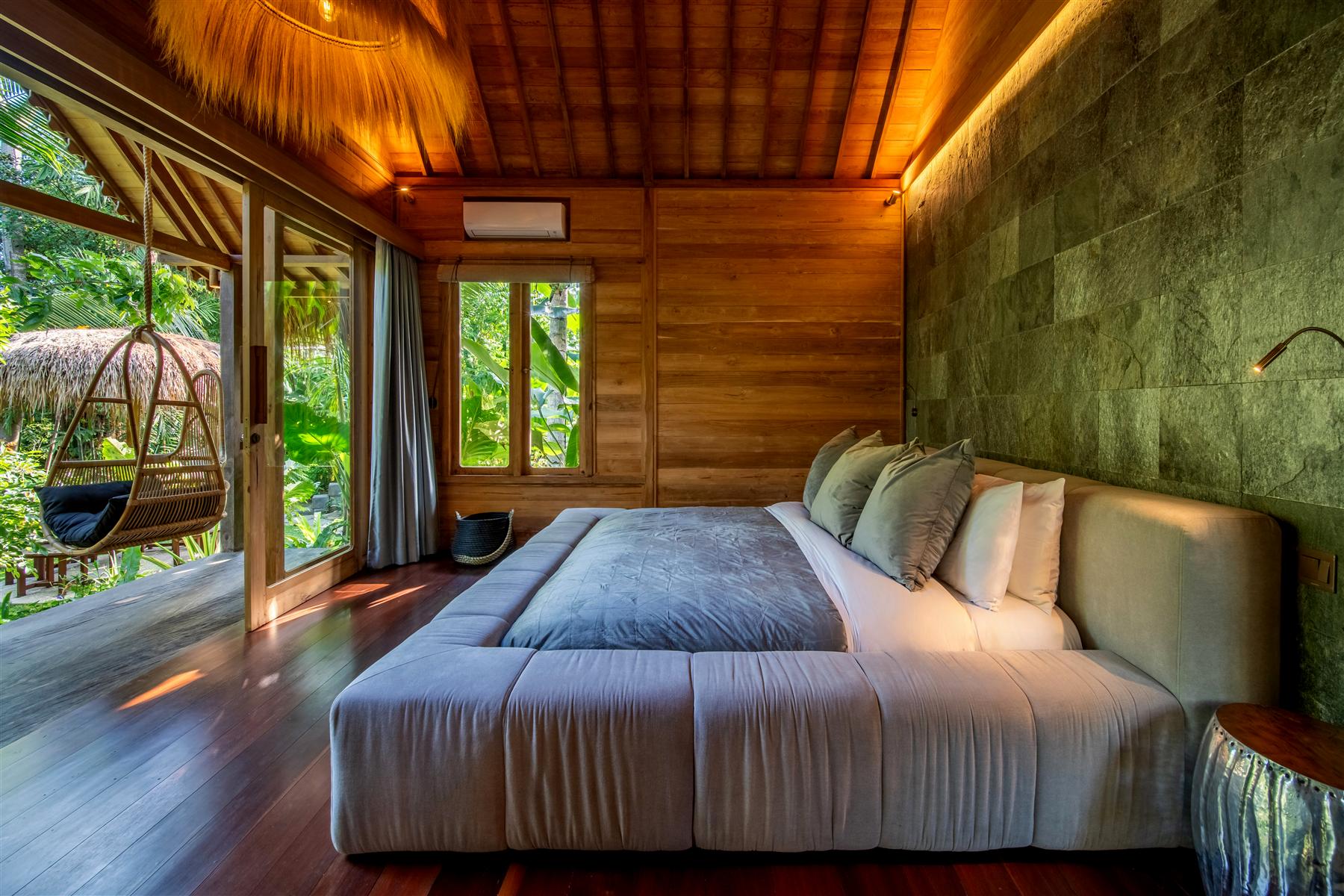 Bedroom at The Seed Bali with wooden walls, stone accent wall, and hanging rattan chair overlooking tropical garden