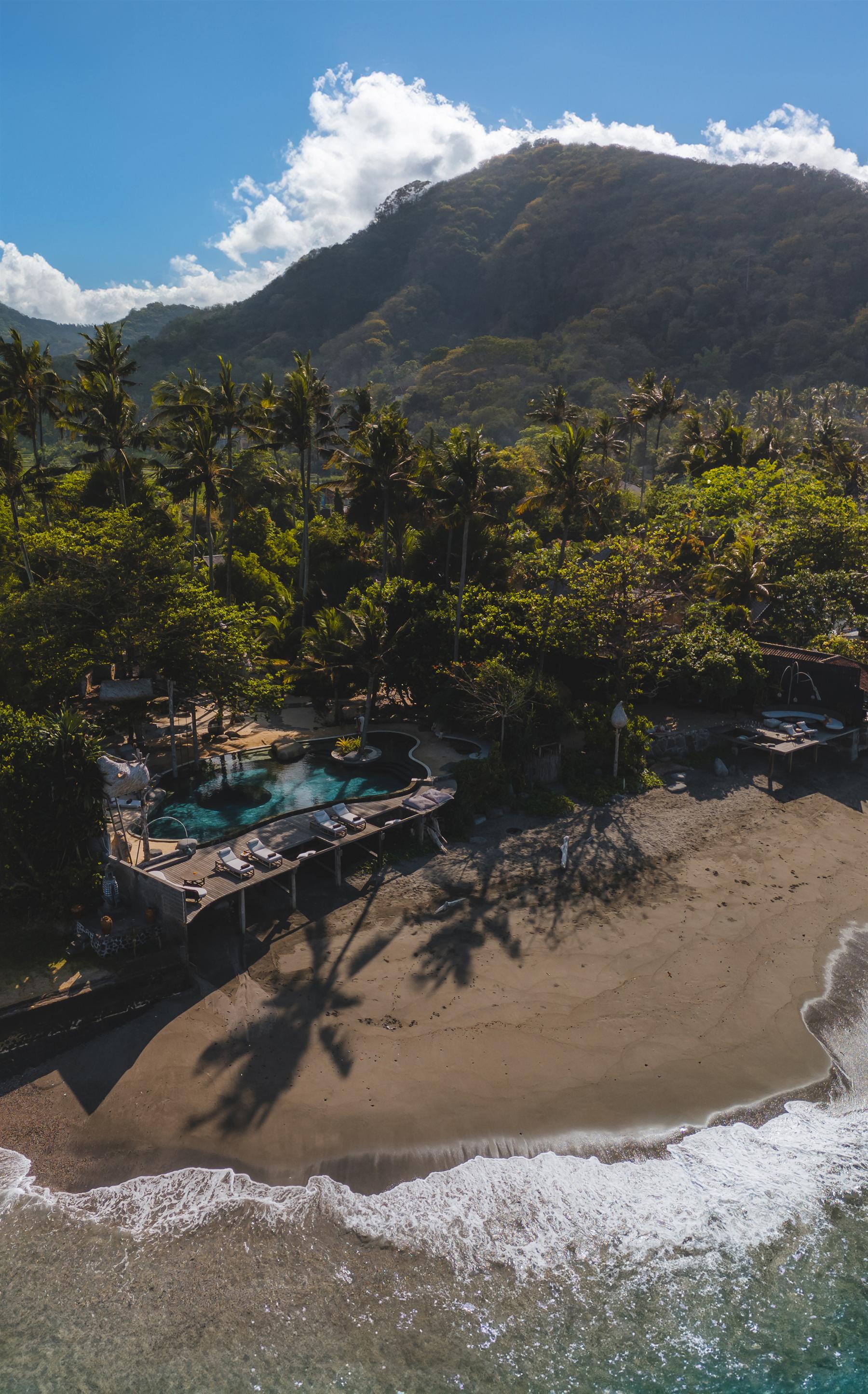 Aerial view of The Seed resort beachfront pool, palm trees, and forested mountain in Candidasa, Bali