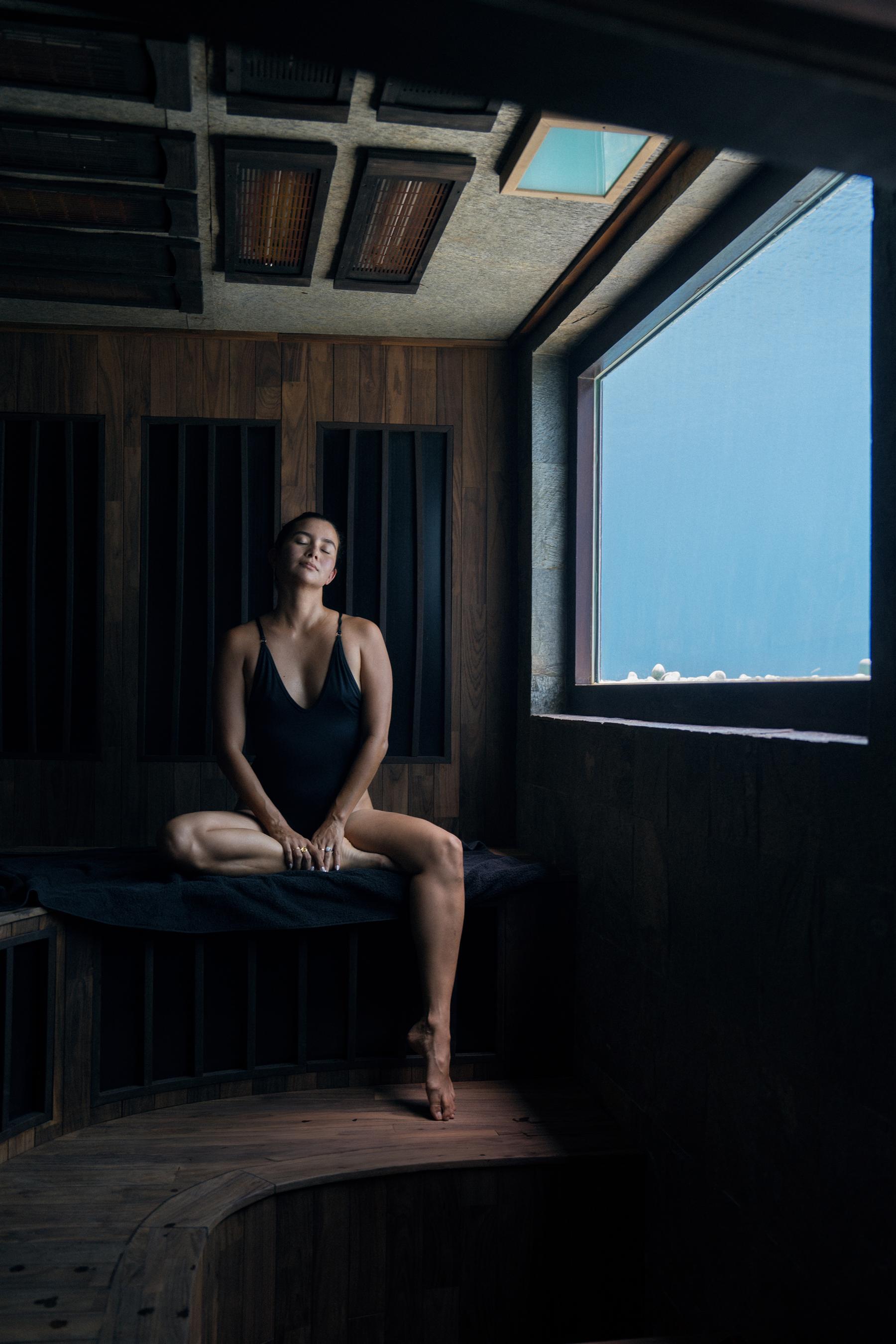 Woman in a dark swimsuit seated inside the wood-paneled sauna at The Seed, Bali, beside a large ocean-view window