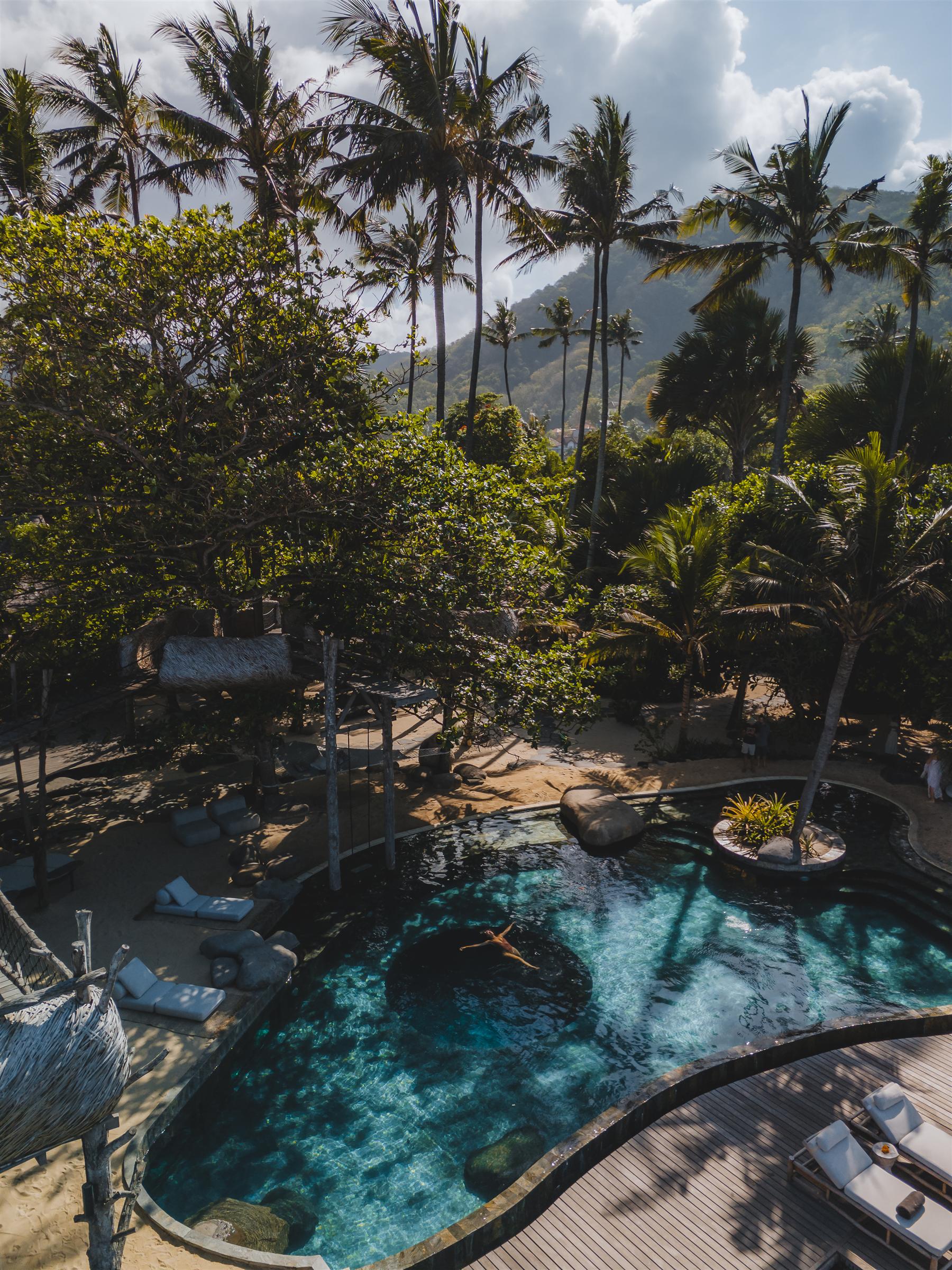 Aerial view of a swimmer in the curved pool at The Seed resort in Candidasa, Bali, surrounded by palm trees and mountains