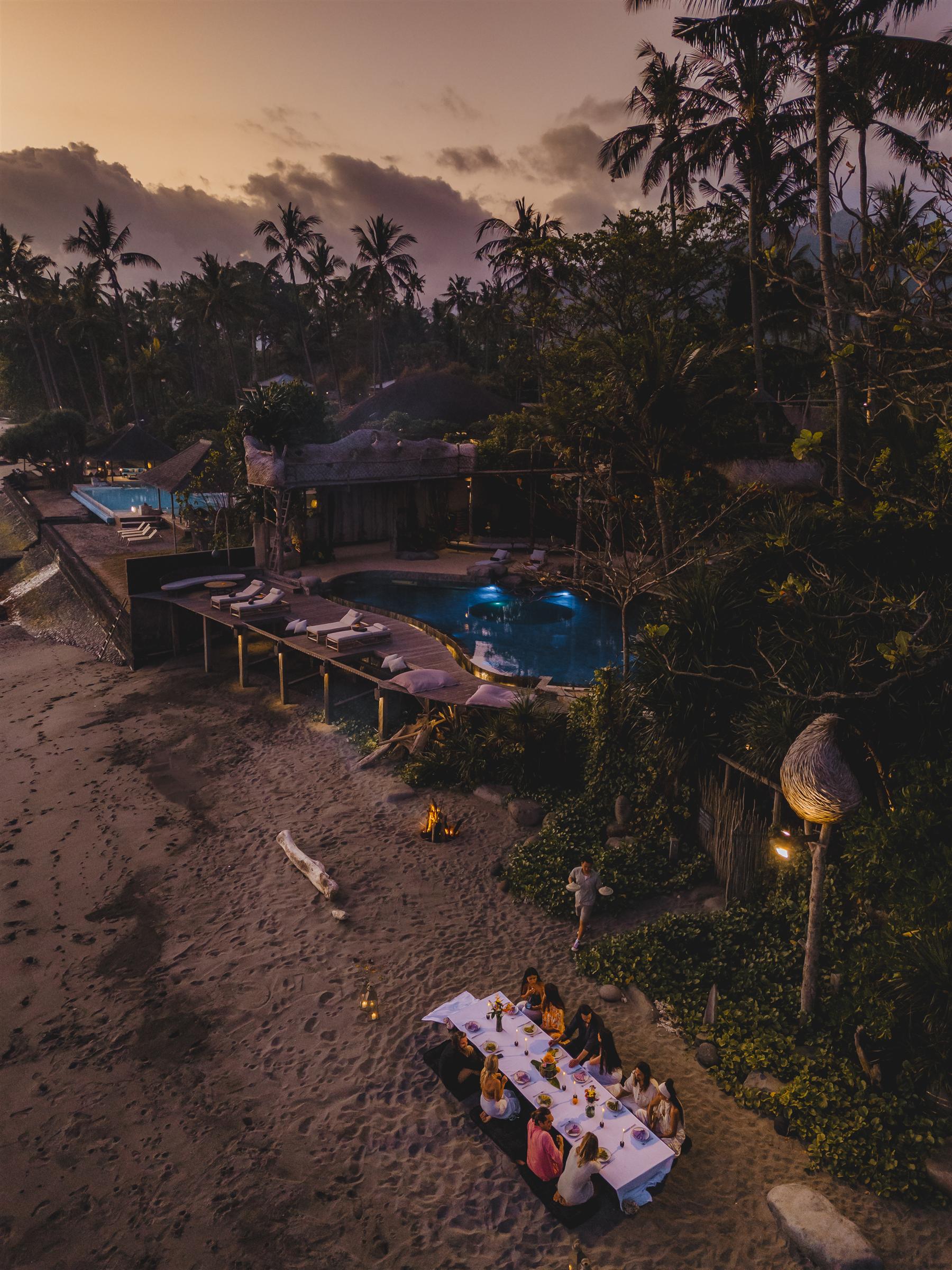 Aerial view of guests dining on the beach at The Seed resort in Candidasa, Bali, at dusk beside a lit pool and palm trees