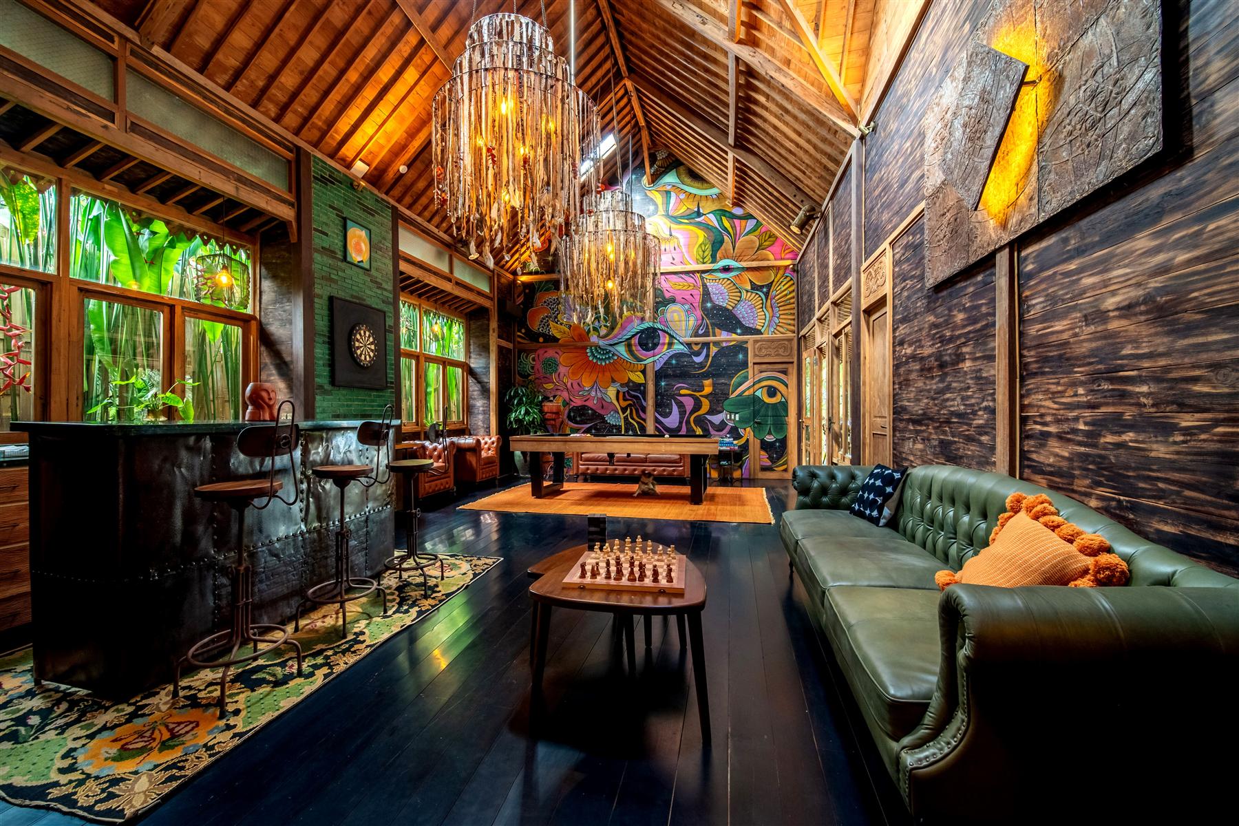 The Seed Bali lounge with vaulted wooden ceiling, chandeliers, green leather sofa, bar, and colorful mural